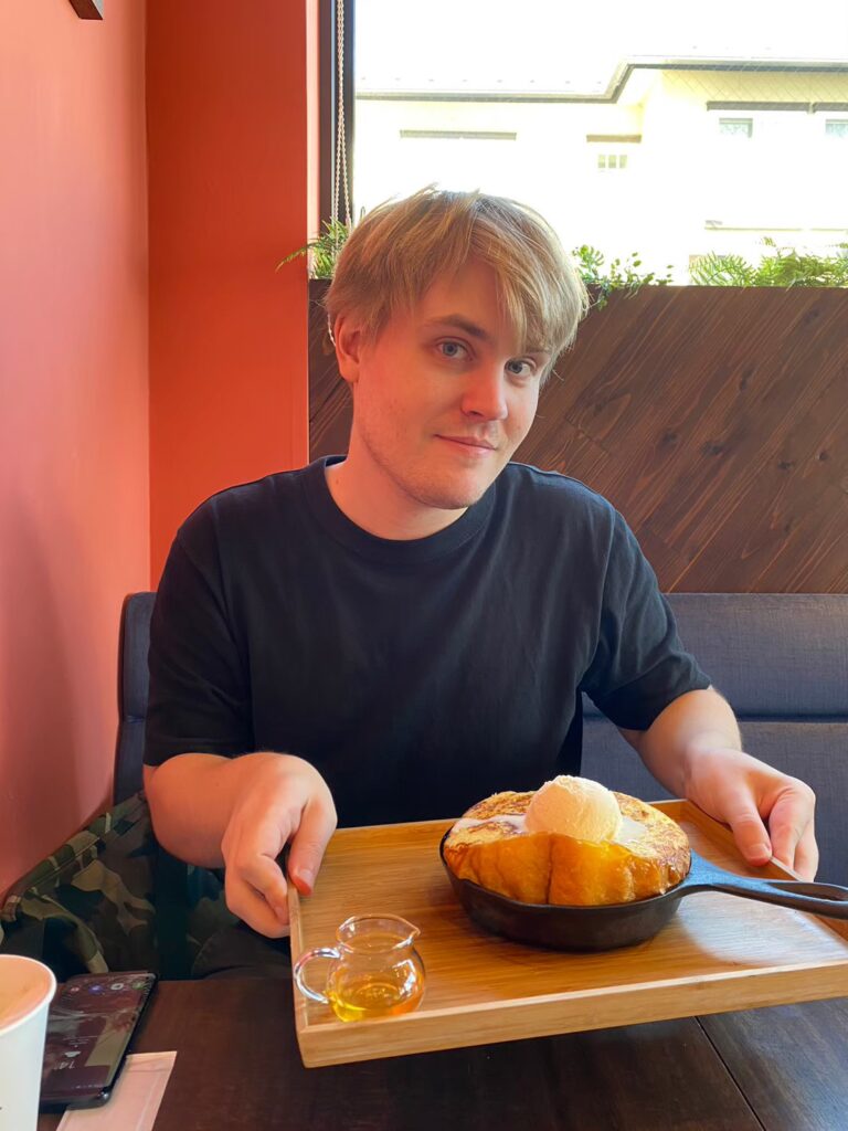 A young man with blonde hair sits at a table in a cafe or restaurant, holding a wooden tray with a cast iron skillet dessert topped with ice cream and a pitcher of syrup. The background includes a red wall, a wooden panel, and a window. This image does not relate to the provided keywords related to expat career guide, advice, Daijob, New Zealand, overseas, software engineer, N2, RPO, consultant, dream job Japan, work in Japan, or jobs in Japan for foreigners.