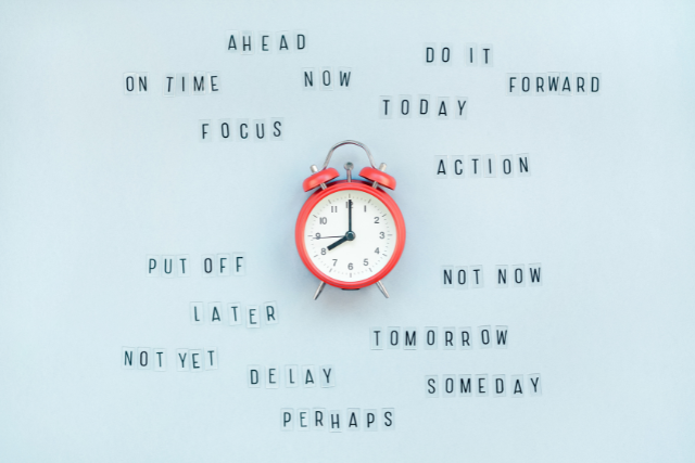 A red alarm clock displaying the time around 8:00 on a light blue background, surrounded by words like AHEAD, ON TIME, FOCUS, PUT OFF, LATER, NOT YET, DELAY, PERHAPS, NOW, DO IT, TODAY, FORWARD, ACTION, NOT NOW, TOMORROW, SOMEDAY. This image conveys the concept of time management.